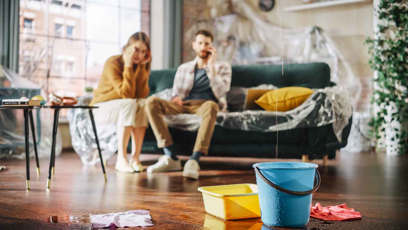Roof is Leaking or Pipe Rupture at Home: Panicing Couple In Despair Sitting on a Sofa Watching How Water Drips into Buckets in their Living Room.